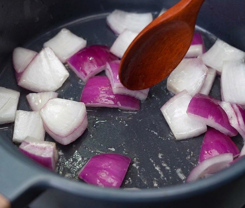 Rote Zwiebelstücke werden mit einem Kochlöffel in einem dunklen Topf bei mittlerer Hitze angeschwitzt.