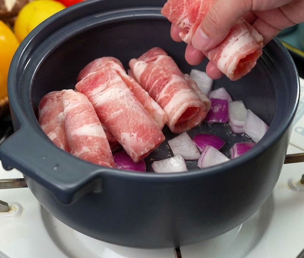 Eine Hand legt dünne Scheiben rohes Rindfleisch zu den angebratenen Zwiebeln in den Topf.