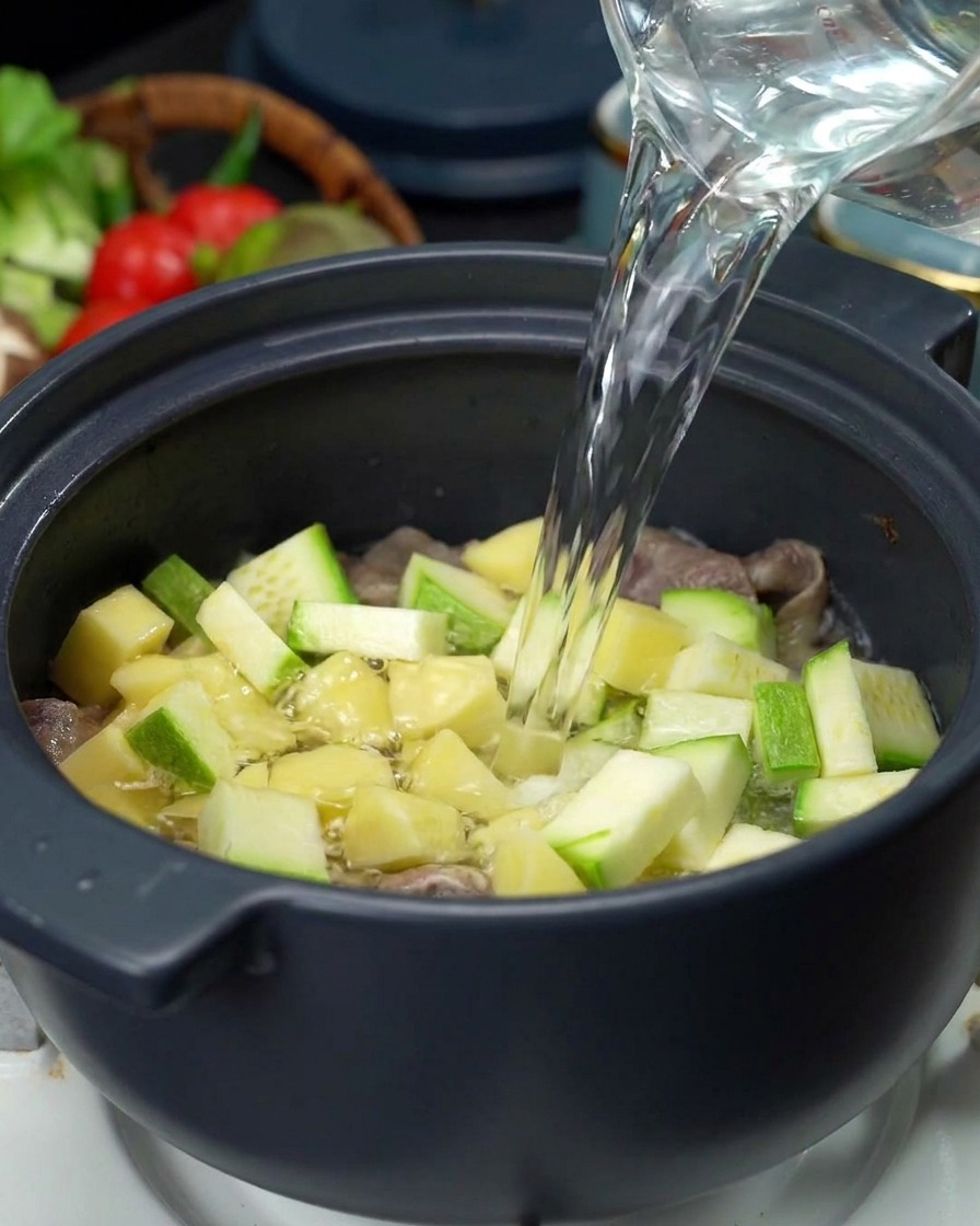 Klares Wasser wird aus einem Glaskrug in den Topf mit Fleisch, Kartoffeln und Zucchini gegossen.
