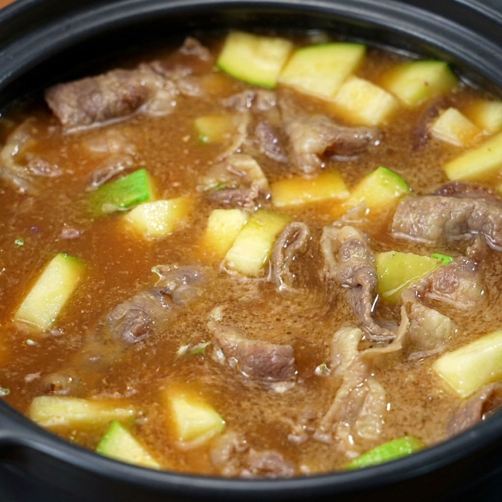 Die Sojabohnenpaste löst sich im Topf auf und färbt die Flüssigkeit in eine trübe, braune Brühe mit Fleisch und Zucchini.