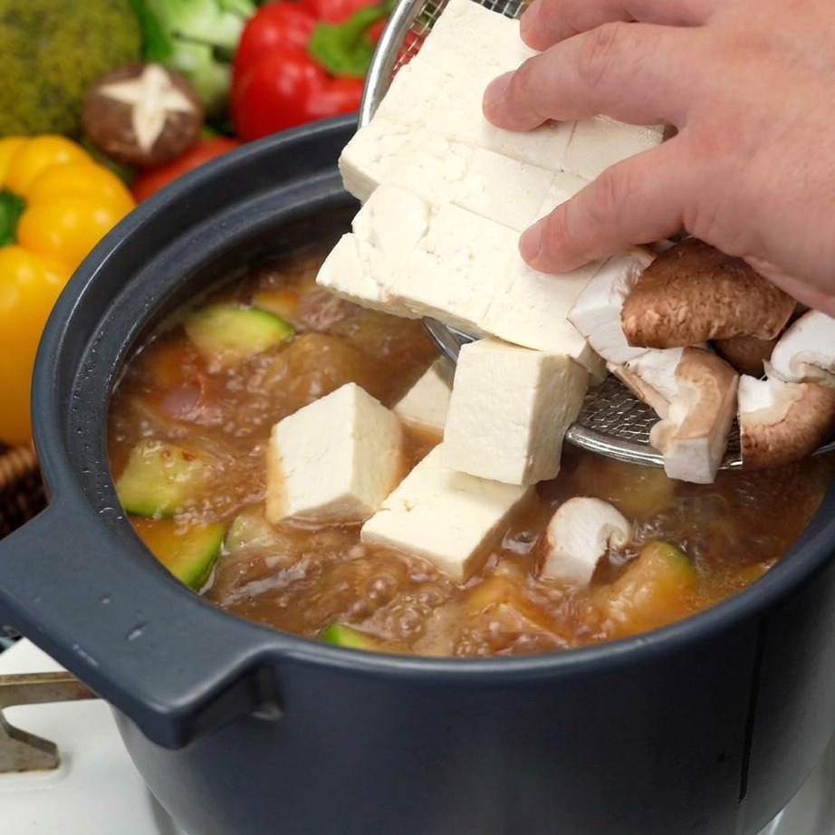 Frische Tofuwürfel und geschnittene Shiitake-Pilze werden vorsichtig in den kochenden Eintopf gegeben.