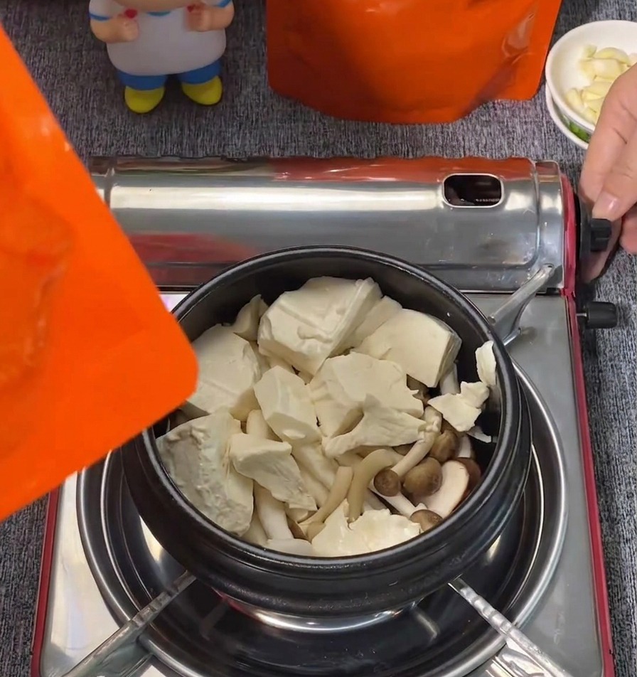 Große Blöcke aus weißem Seidentofu und Shimeji-Pilze sind in einem schwarzen Kochtopf angerichtet.
