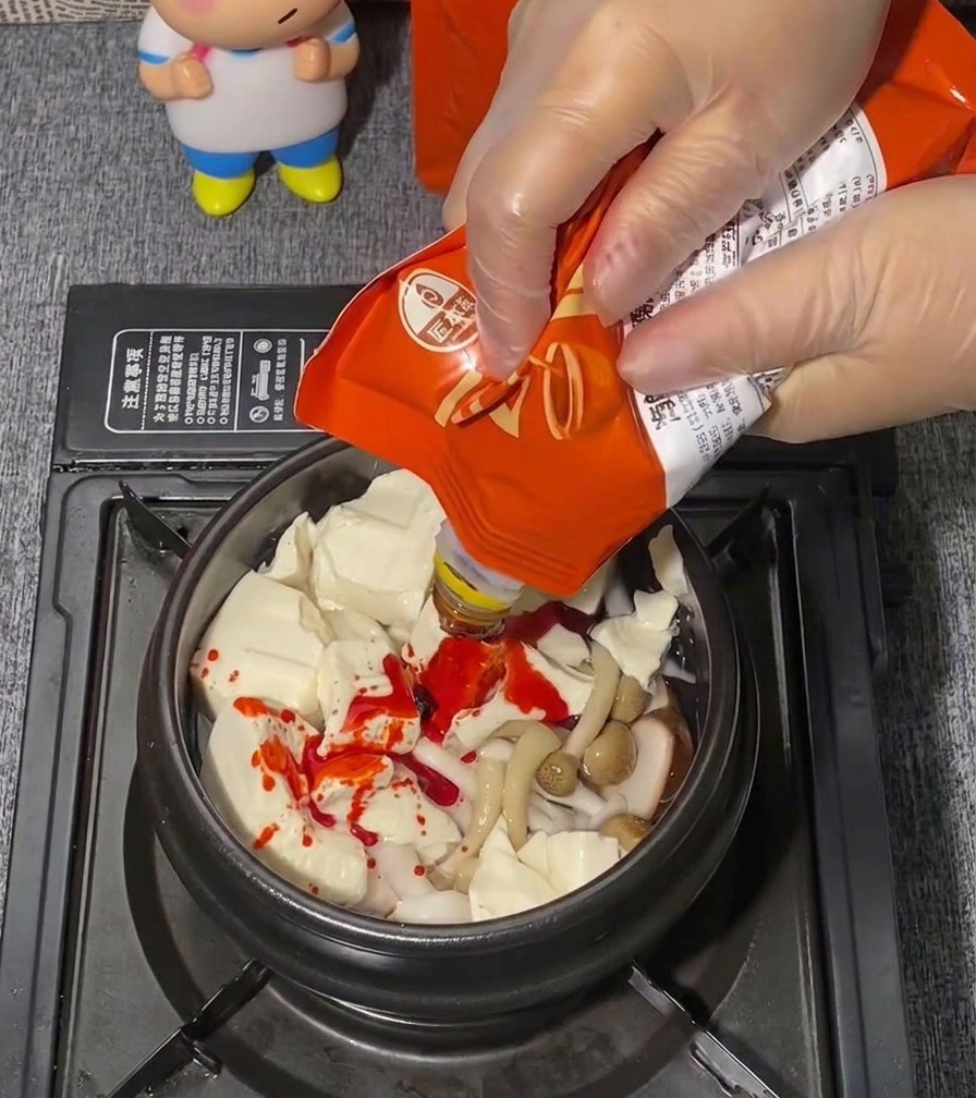 Eine Hand in einem transparenten Plastikhandschuh drückt leuchtend rote Würzpaste aus einem Beutel auf ein Bett aus Seidentofu.