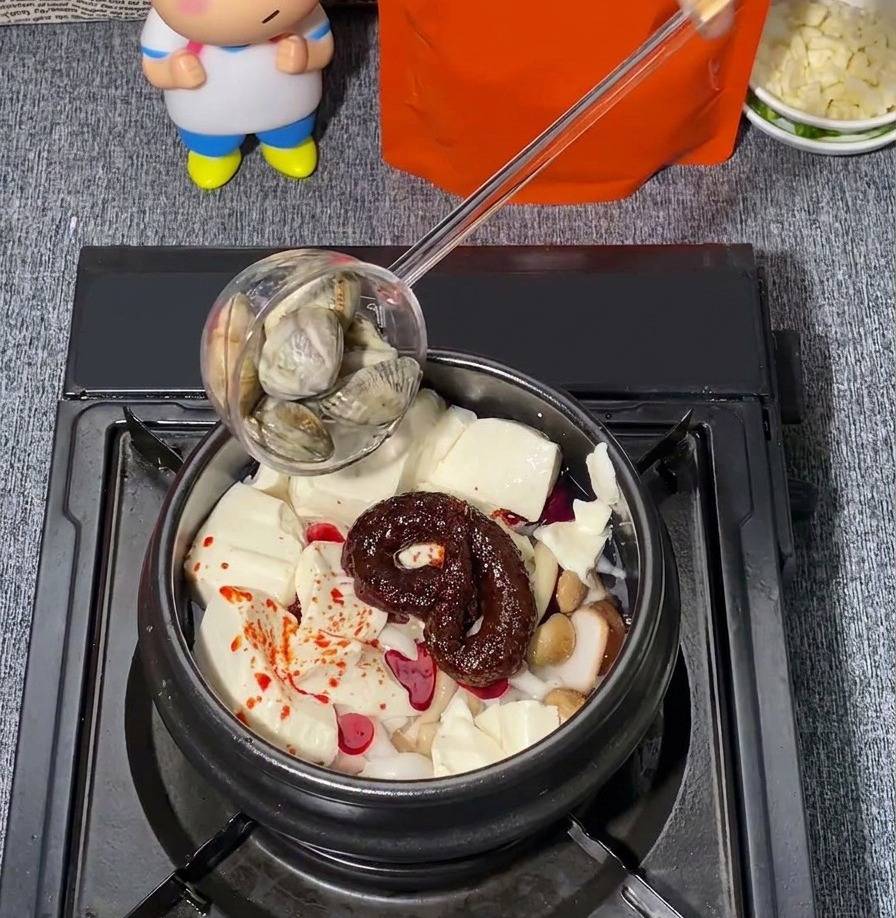 Frische Venusmuscheln werden mit einer Kelle in einen Steintopf mit Seidentofu, Pilzen und rotem Chili-Gewürz gegeben.
