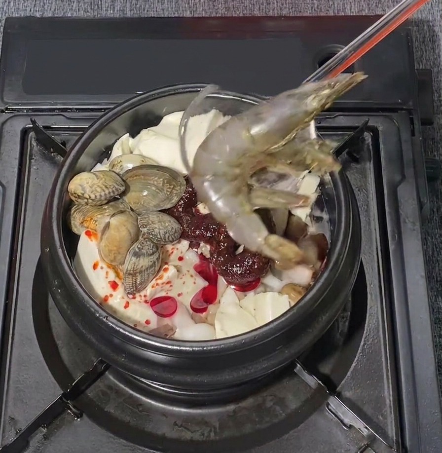 Zwei große rohe Garnelen werden vorsichtig in einen brodelnden Topf mit koreanischem Seidentofu-Eintopf gelegt.