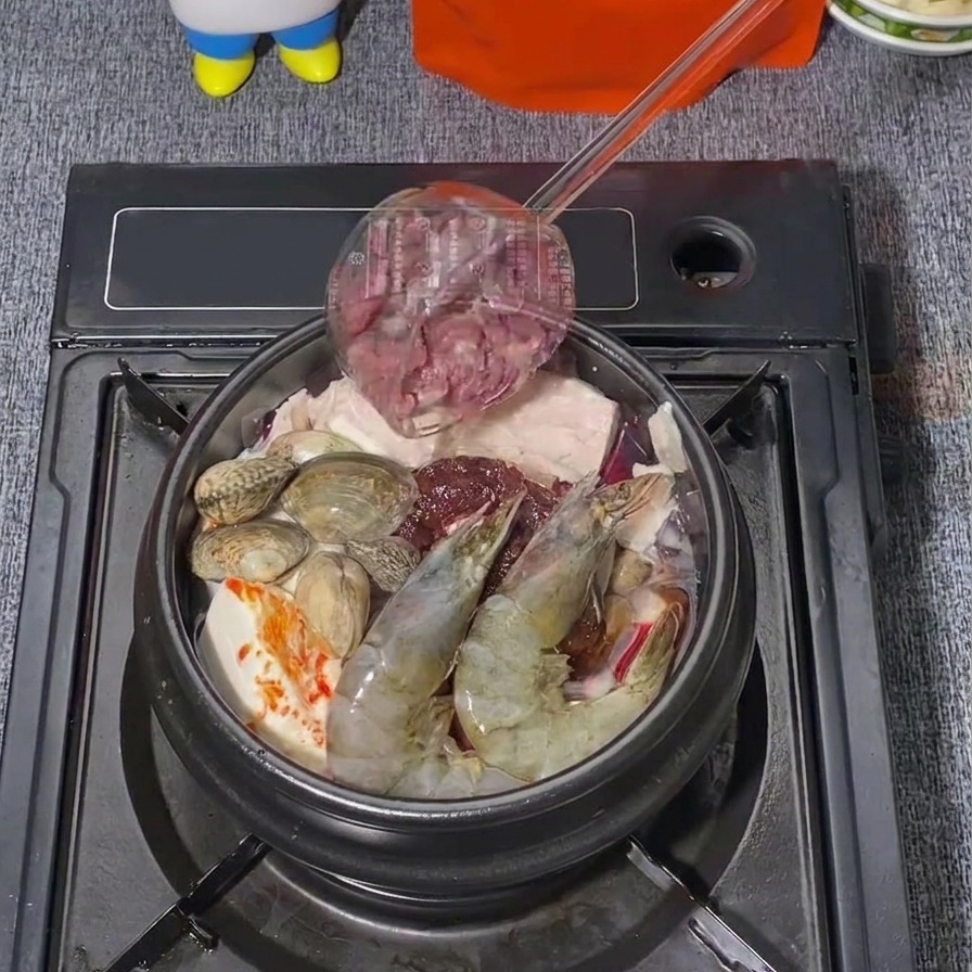 Dünne Scheiben rohes Rindfleisch werden mit einer Kelle in einen koreanischen Eintopf gegeben, der bereits Garnelen und Muscheln enthält.