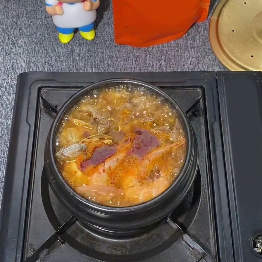 Ein brodelnder Topf mit koreanischem Seidentofu-Eintopf mit einer kräftig roten Brühe, sichtbaren Muscheln und aufsteigendem Dampf.