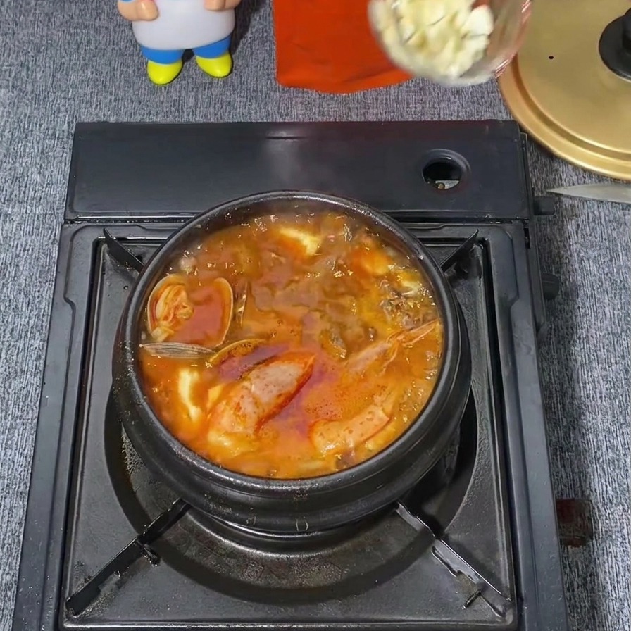 Eine Kelle gibt einen Löffel gehackten weißen Knoblauch in die Mitte eines kochenden, scharf-roten koreanischen Eintopfs.