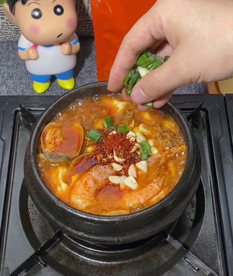 Eine Hand streut eine Portion gehackte grüne Frühlingszwiebeln in einen brodelnden schwarzen Steintopf mit koreanischem Eintopf, der mit Garnelen, Muscheln und Knoblauch gefüllt ist.