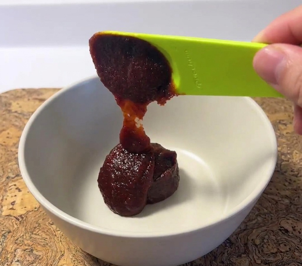 Ein Klecks dicke, dunkelrote koreanische Chilipaste wird mit einem grünen Löffel in eine weiße Keramikschüssel gegeben