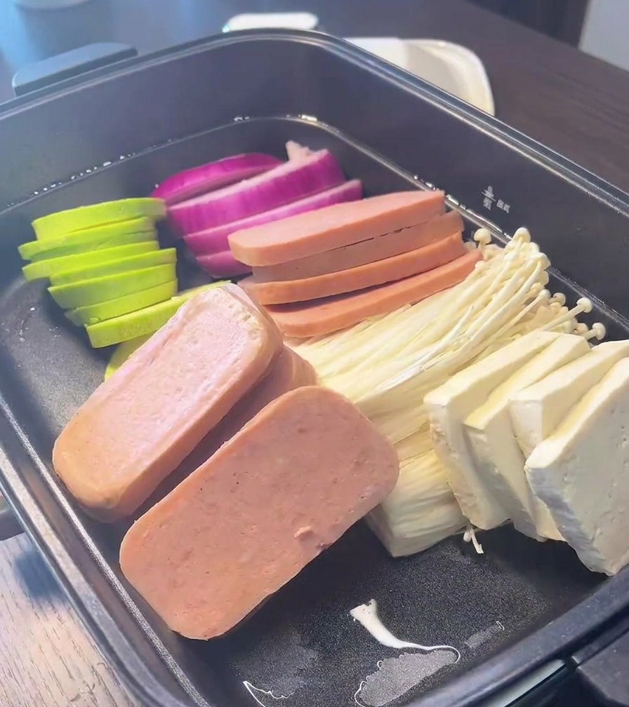Ein schwarzer Kochtopf, gefüllt mit ordentlich arrangierten Abschnitten aus roten Zwiebeln, grüner Zucchini, Dosenfleisch, Enoki-Pilzen und Tofublöcken.