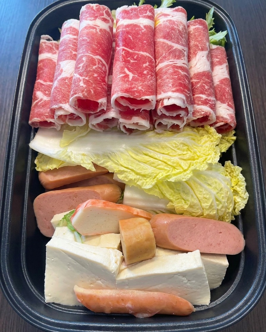 Eine Hand legt mehrere Röllchen aus leuchtend rotem, dünn geschnittenem, marmoriertem Rindfleisch auf die bunten Zutaten im Topf.
