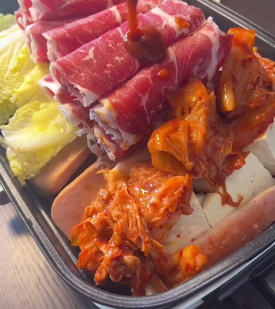Ein Haufen leuchtend rotes, fermentiertes Kimchi liegt in der Mitte eines mit Fleisch und Gemüse gefüllten Topfes.