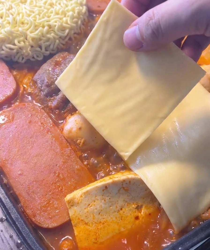 Eine Hand legt eine quadratische Scheibe gelben Käse auf die Instant-Nudeln in einem brodelnden roten Eintopf.
