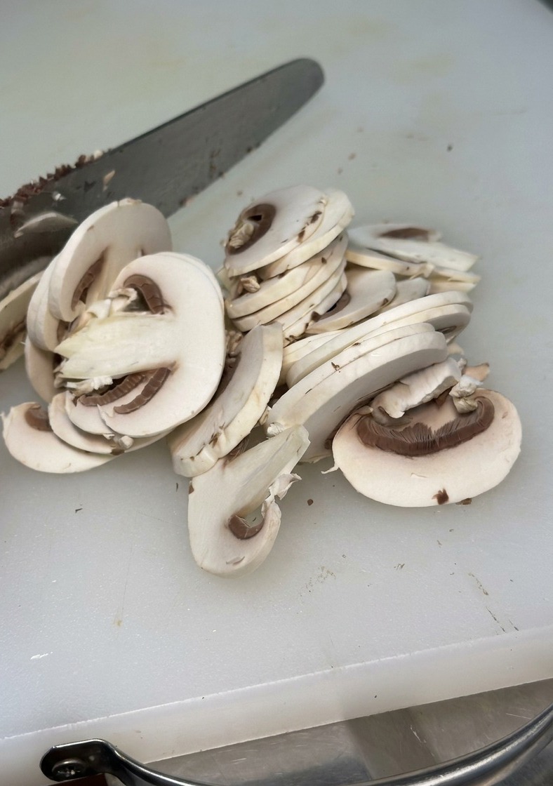 Nahaufnahme eines weißen Champignons, der mit einem Metallmesser auf einem weißen Schneidebrett in dünne, gleichmäßige Scheiben geschnitten wird.