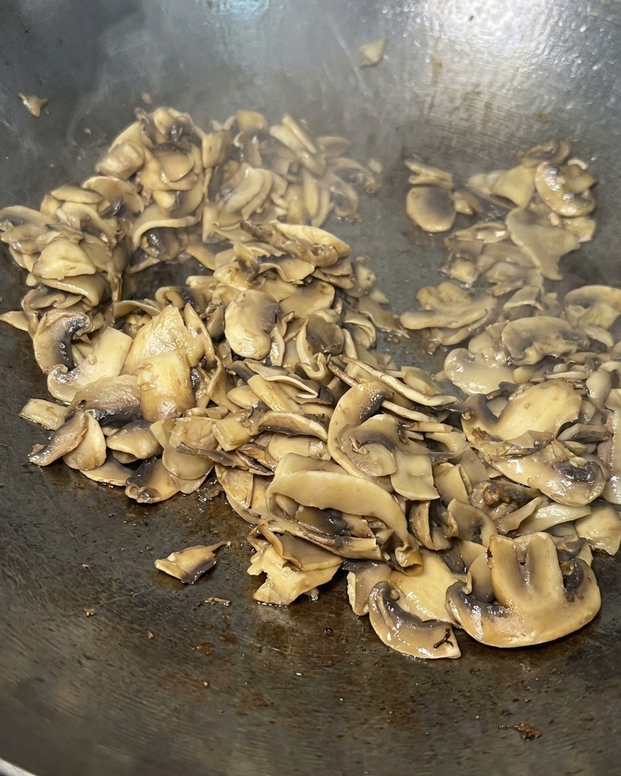 Angebratene Champignonscheiben in einer Pfanne, goldbraun und weitgehend trocken gegart.