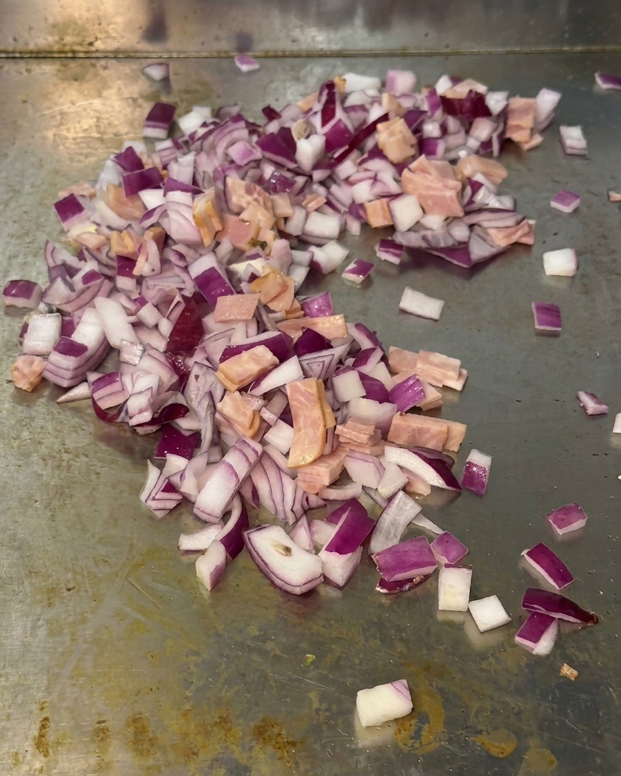 Eine Mischung aus roten Zwiebelwürfeln und kleinen Speckstücken, die zusammen auf einer Metallgrillfläche angebraten werden.