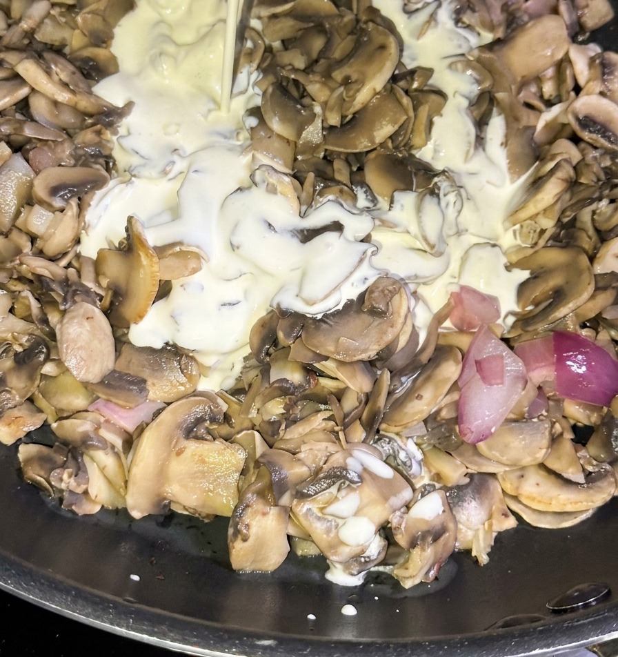 Ein Strahl weißer Schlagsahne wird aus einem blauen Karton in eine Pfanne mit angebratenen Champignons und roten Zwiebelwürfeln gegossen.