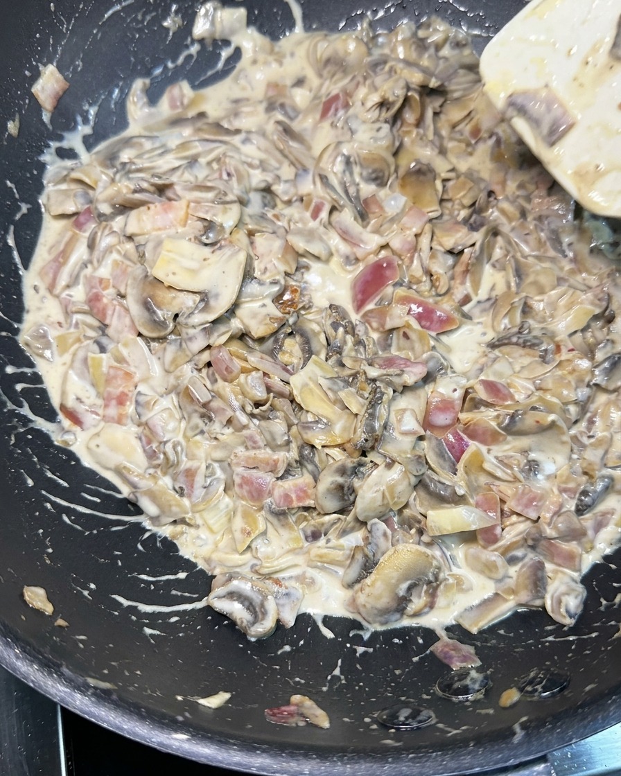 Ein weißer Pfannenwender rührt eine dicke, cremige Sauce mit Champignonscheiben, roten Zwiebelwürfeln und Speckstücken in einer schwarzen Pfanne.