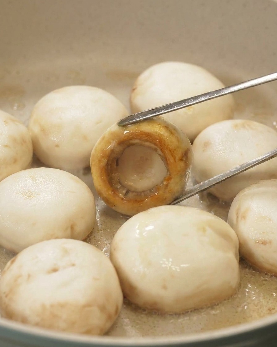 Eine Zange wendet einen Pilz in der Pfanne und legt eine perfekt karamellisierte, goldbraune Oberfläche frei