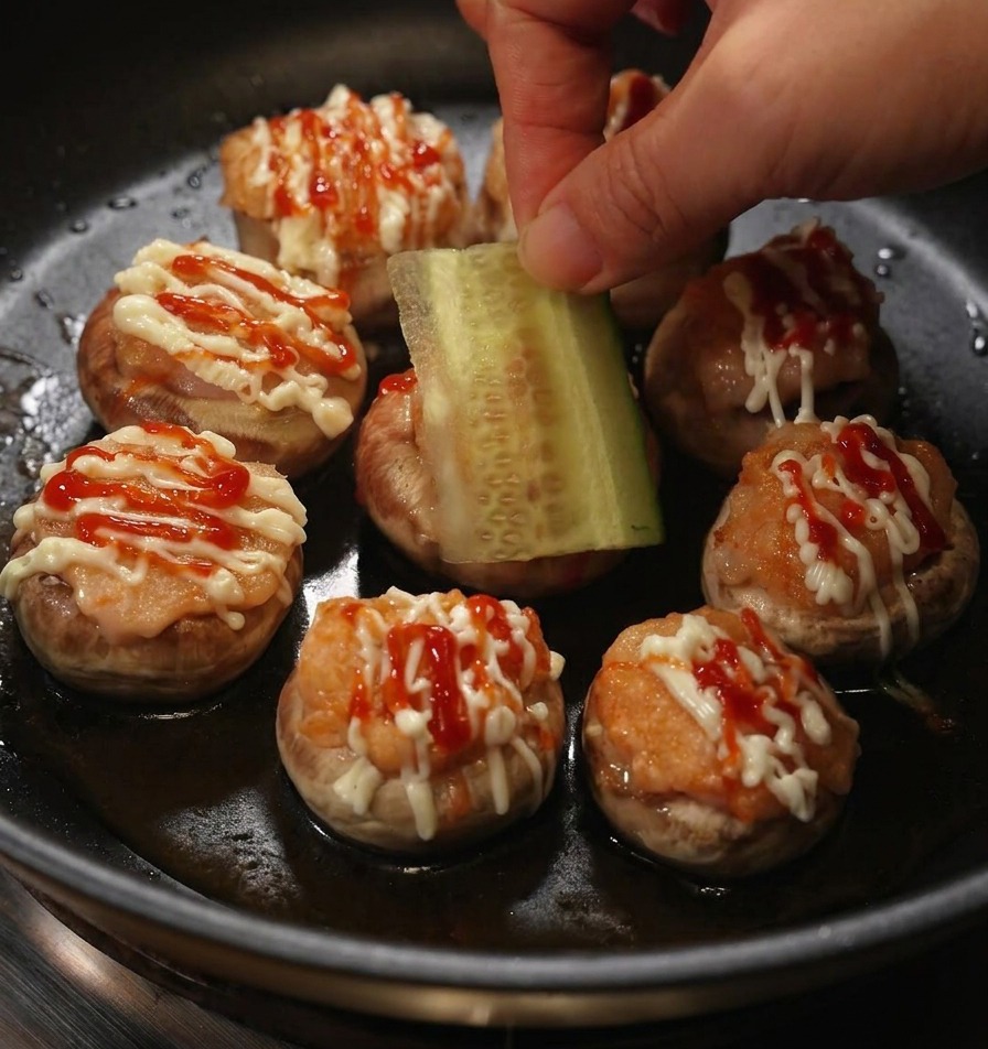 Eine Hand legt eine rechteckige Scheibe frische Gurke auf einen gefüllten Pilz-Burger mit Mayonnaise und Ketchup