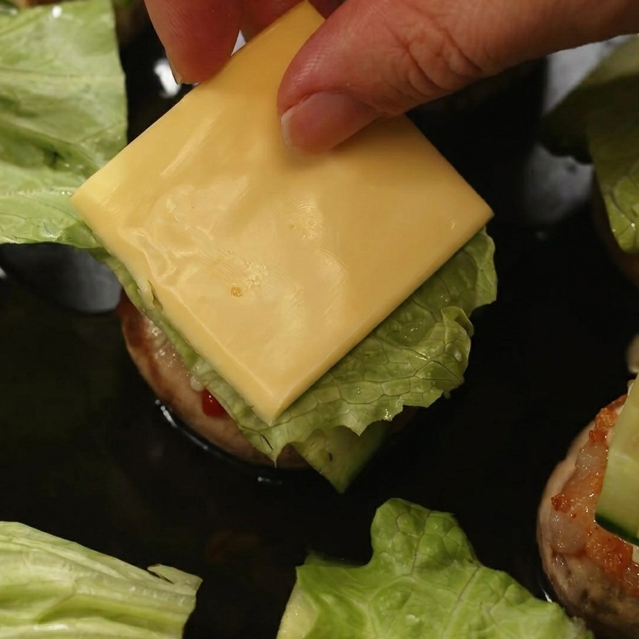 Eine Hand legt eine quadratische Scheibe gelben Käse auf Salat und Gurke in einem Pilz-Burger