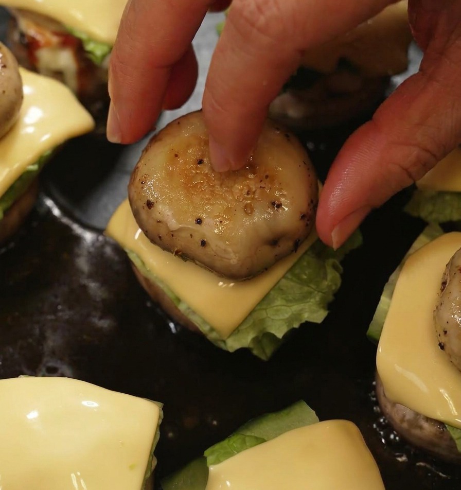 Eine Hand setzt einen gegrillten Pilzkopf auf einen Mini-Burger, um das Schichten in der Pfanne abzuschließen