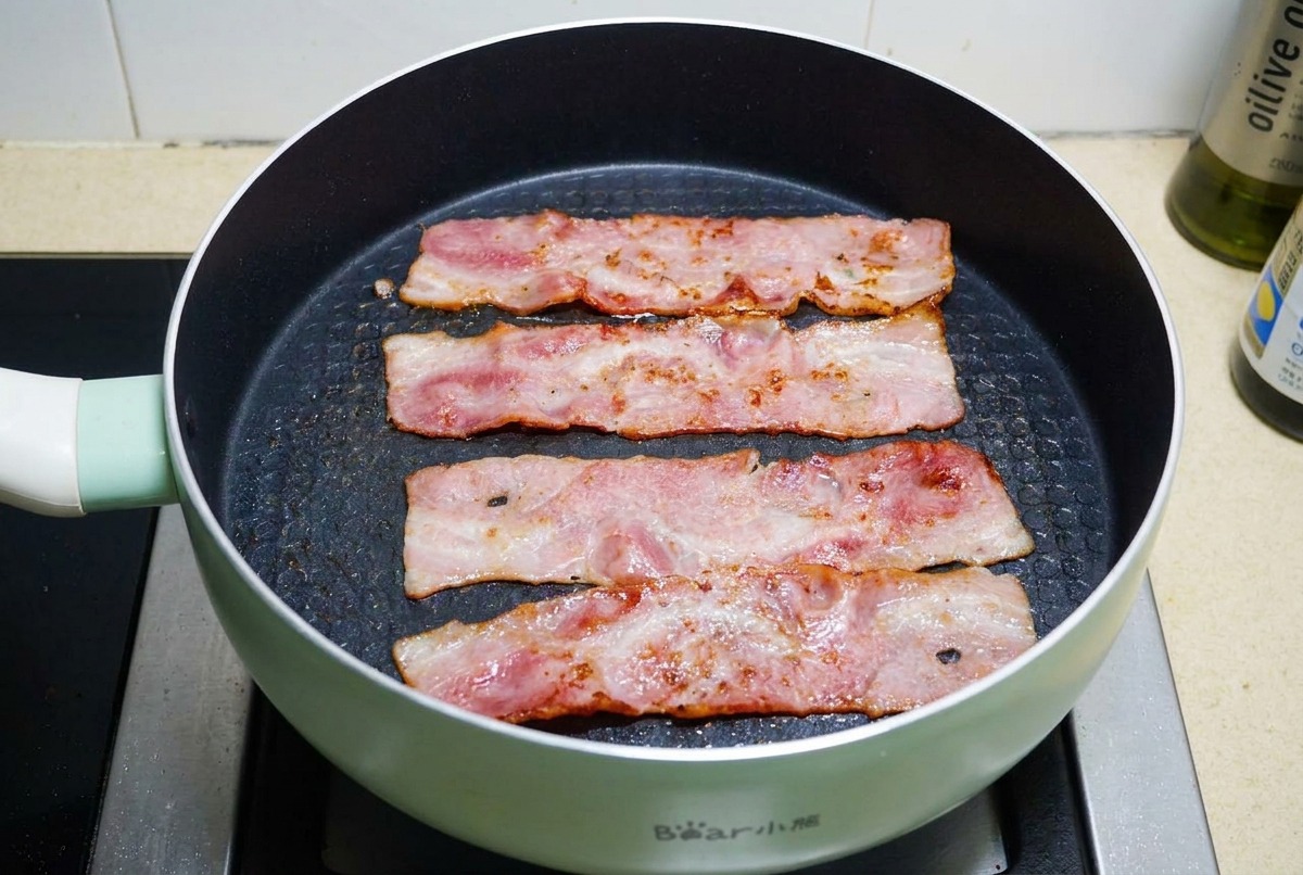 Vier Streifen Bacon braten in einer schwarzen beschichteten Pfanne, bis das Fett blubbert und der Speck goldbraun und knusprig ist.