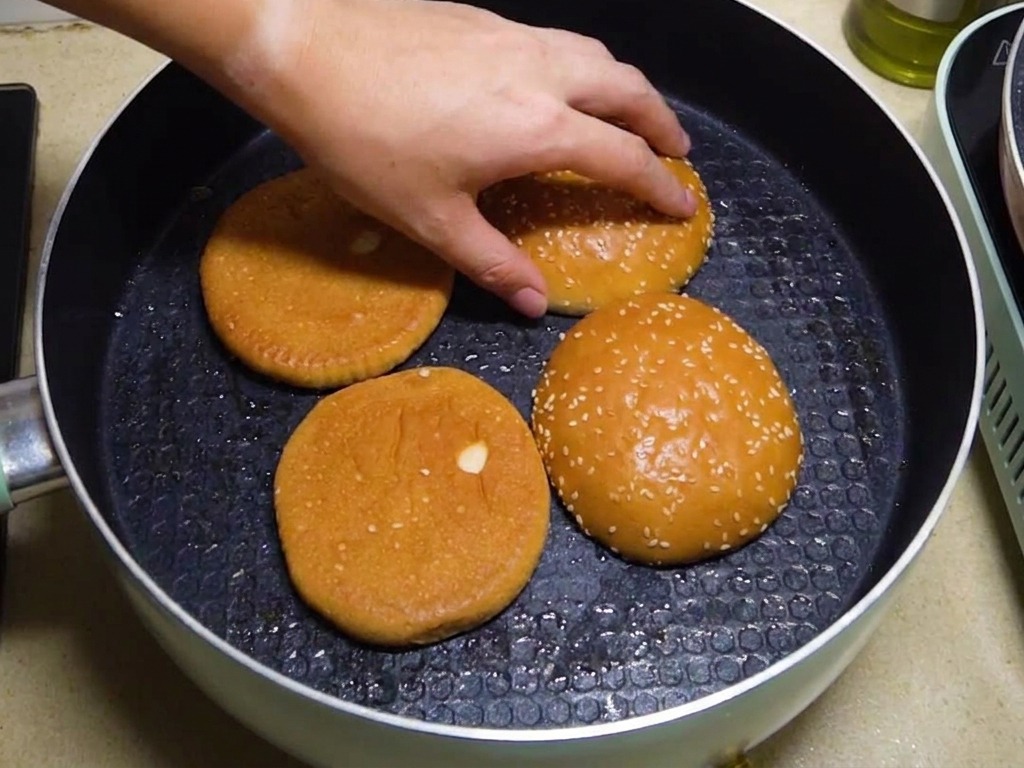 Vier Burgerbrötchen-Hälften werden mit der Schnittfläche nach unten in einer Pfanne getoastet, während eine Hand ihre Position anpasst.