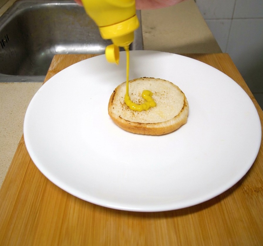 Eine Hand drückt leuchtend gelben Senf aus einer Plastikflasche auf ein getoastetes Burgerbrötchen auf einem weißen Teller.