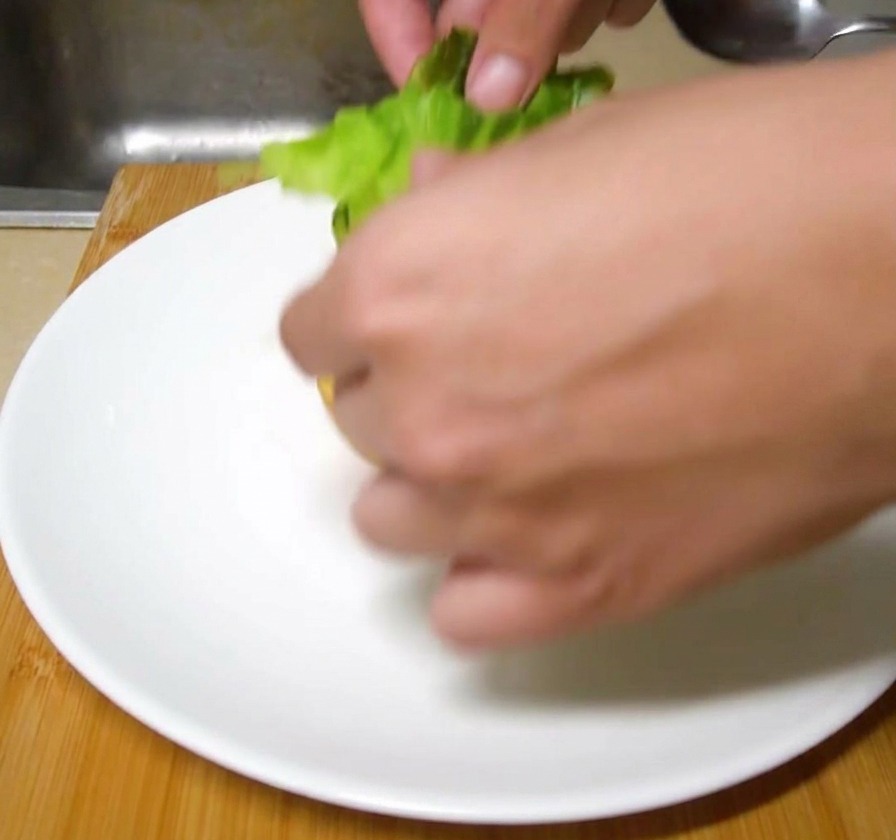 Frische grüne Salatblätter werden von Hand auf ein getoastetes Burgerbrötchen auf einem weißen Keramikteller gelegt.
