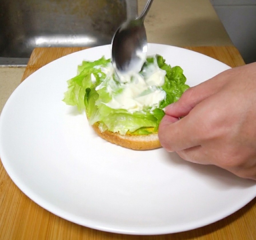 Ein Metalllöffel verstreicht einen Klecks weiße Mayonnaise auf frischen grünen Salatblättern eines Burgers.