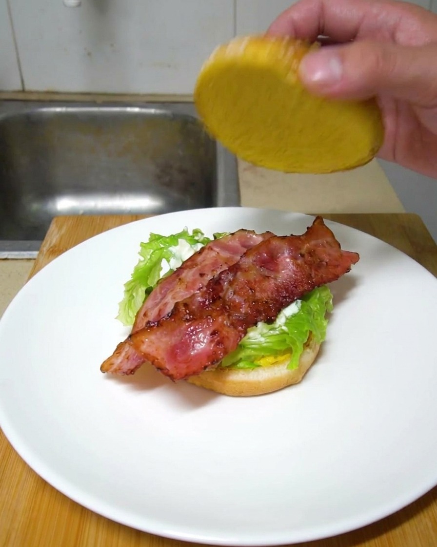 Eine Hand setzt die obere Brötchenhälfte auf einen fertig geschichteten Burger mit Salat und knusprigen Baconstreifen.
