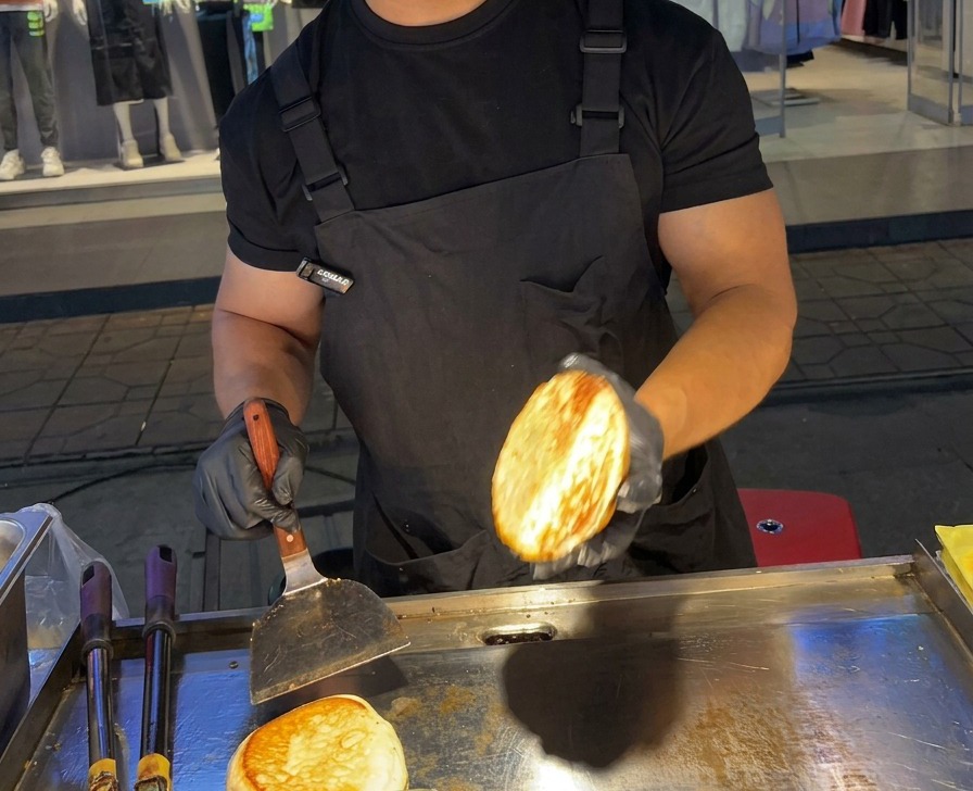 Eine Person hält eine perfekt geröstete, goldbraune Burger-Brötchenhälfte über eine flache Edelstahl-Grillplatte.