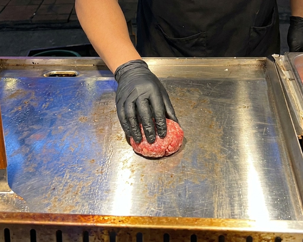Eine Hand in einem schwarzen Handschuh drückt eine Kugel aus rohem Rinderhackfleisch mit einer Metallpresse auf einer heißen Grillplatte flach.