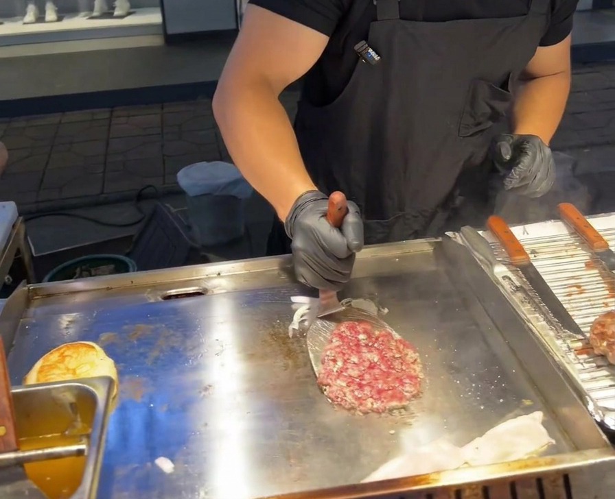 Ein Metall-Pfannenwender wendet ein Rinder-Patty auf einer Grillplatte direkt über einem Haufen gegrillter roter Zwiebeln.