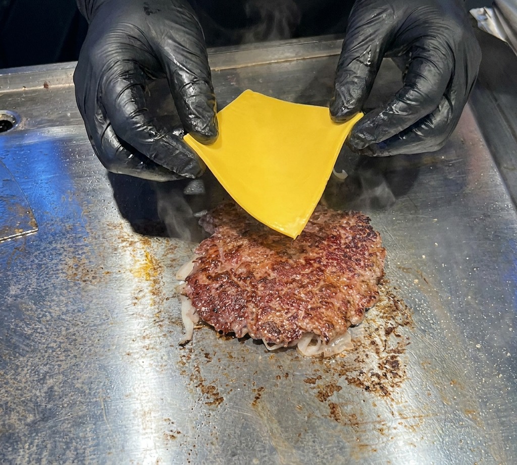 Nahaufnahme von Händen in schwarzen Handschuhen, die eine Scheibe gelben Cheddar-Käse auf ein brutzelndes, braunes Rinder-Patty legen.