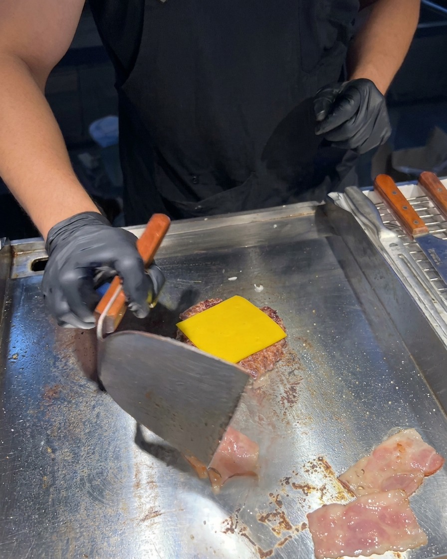 Ein Metall-Pfannenwender wendet brutzelnde Baconstreifen auf einer Grillplatte neben einem Patty mit geschmolzenem Käse.
