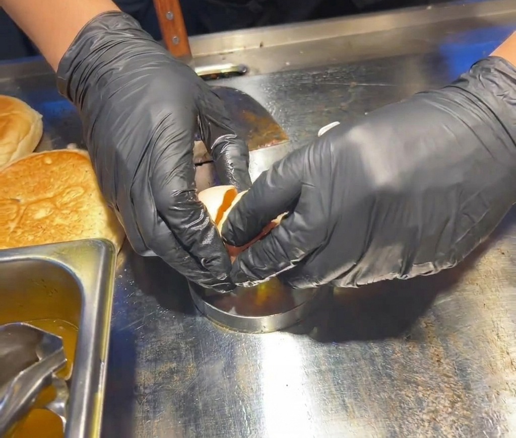 Hände in schwarzen Handschuhen schlagen ein frisches Ei in einen silbernen, runden Metallring auf einer Edelstahl-Grillfläche.