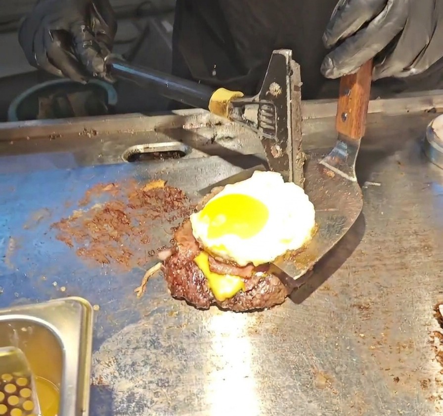 Ein breiter Metall-Pfannenwender hebt einen hohen Burger-Stapel aus Patty, geschmolzenem Käse, Bacon und einem Spiegelei an.