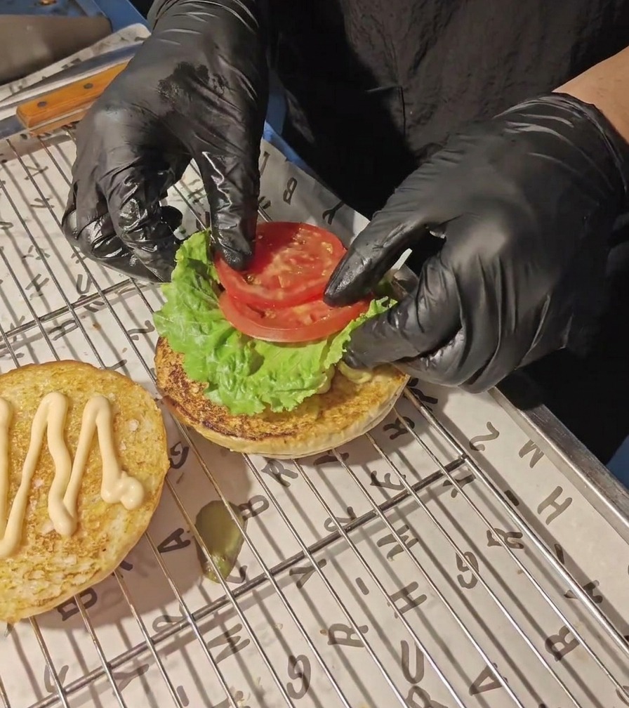 Hände in schwarzen Handschuhen legen frische Tomatenscheiben auf ein grünes Salatblatt auf der unteren Brötchenhälfte.