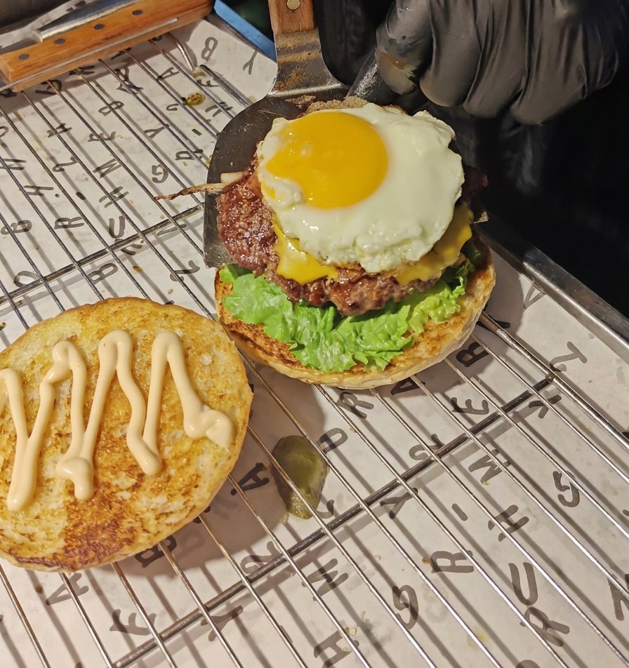 Ein Metall-Pfannenwender setzt ein Rinder-Patty mit geschmolzenem Käse und Spiegelei auf die Salat- und Tomatenschicht.