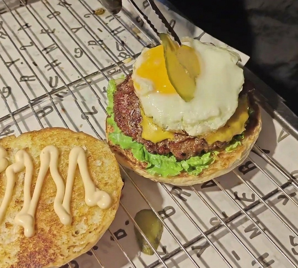 Eine Zange platziert eine dünne Gewürzgurkenscheibe auf einem strahlend gelben Eigelb inmitten eines Burger-Stapels.