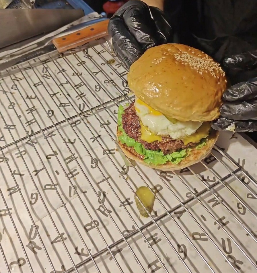 Hände in schwarzen Handschuhen setzen eine goldbraune Brötchenoberseite auf einen fertig geschichteten Ei- und Bacon-Burger.