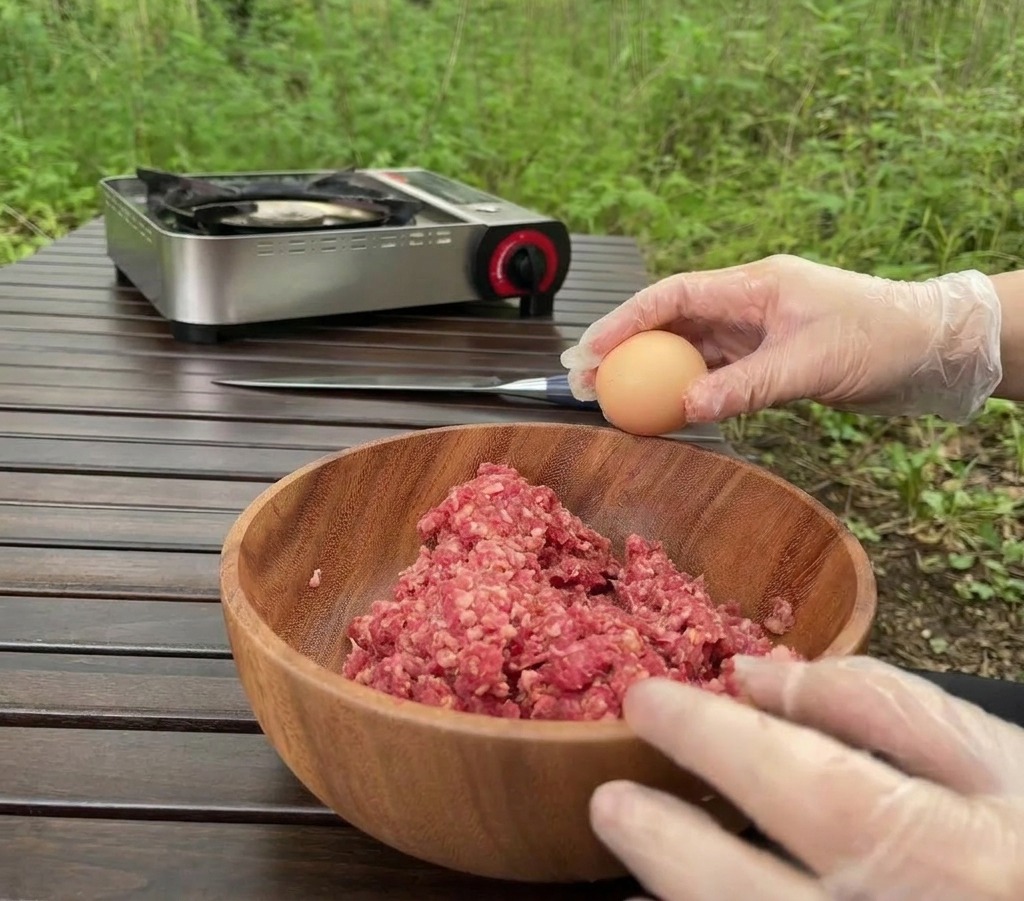 Eine Person mit durchsichtigen Handschuhen schlägt im Freien über einer Holzschüssel voll Rinderhackfleisch ein braunes Ei auf.