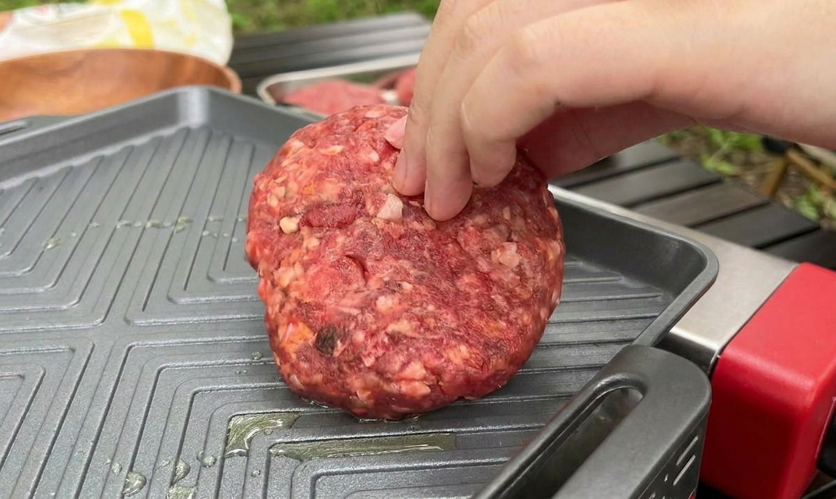 Eine Hand legt ein dickes, rohes Rindfleisch-Patty auf eine heiße, geriffelte Grillpfanne.
