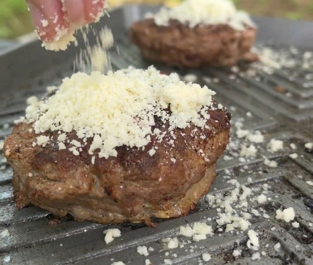 Nahaufnahme: Geriebener weißer Käse wird auf ein gebräuntes Rindfleisch-Patty auf einer heißen Grillpfanne gestreut.
