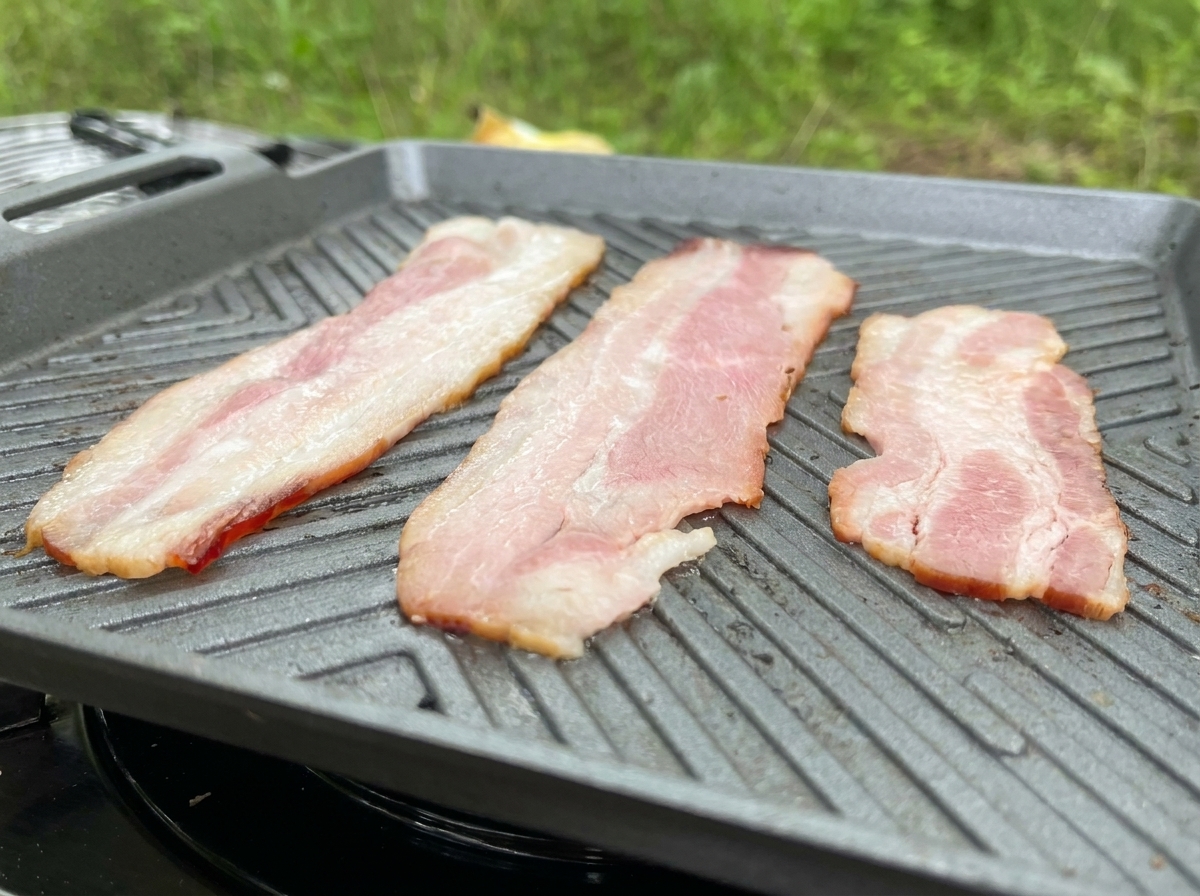 Drei lange Streifen Bacon braten auf einer grauen geriffelten Grillpfanne; das Fett brät deutlich aus.