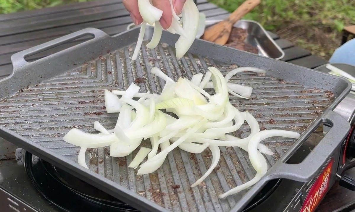 Dünn geschnittene weiße Zwiebeln werden neben dem Fleisch auf die heiße Grillpfanne gegeben.