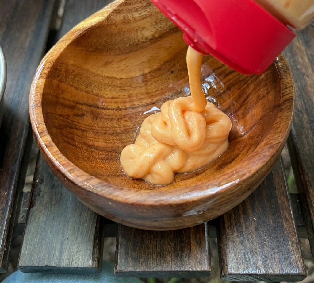 Cremige orange Sauce wird aus einer Flasche mit rotem Deckel in eine kleine Holzschüssel zu anderen Saucenzutaten gedrückt.